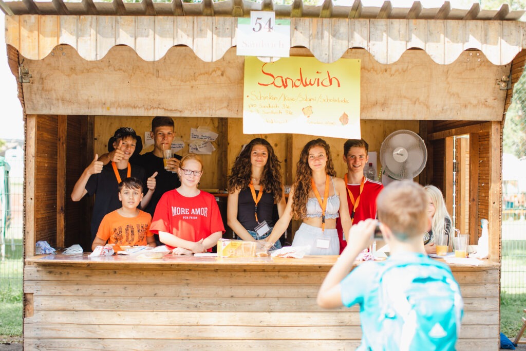 Spielkinder und jugendliche Helfer stehen gemeinsam hinter einem Verkaufsstand in der Spielstadt Bad Kresselnau.